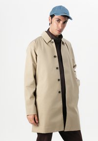 Casaco bege com colarinho clássico, apresentando botões escuros, bolsos laterais e um corte reto. Usado sobre uma peça de roupa castanha e acompanhado de um boné azul.