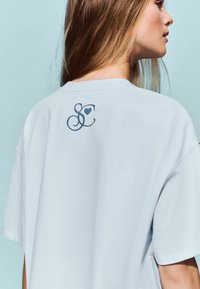 T-shirt en coton bleu clair avec une coupe décontractée, arborant un logo "SC" bleu foncé au dos, des manches courtes et des fentes latérales pour un confort de mouvement.