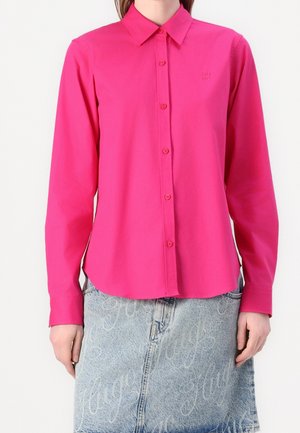Femme portant une chemise rose vif à manches longues boutonnée, associée à une jupe en jean bleu clair avec des motifs de texte blanc en cursive.