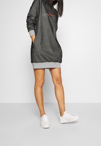 Vestido sudadera con capucha de color gris oscuro y dobladillo gris claro, con un logo naranja. Combinado con zapatillas blancas, tiene bolsillos laterales y mangas largas.