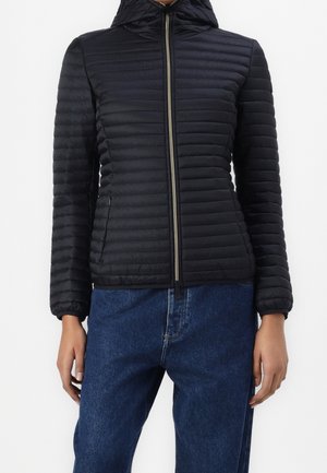 Personne portant une veste matelassée noire à capuche zippée et un jean bleu, debout, les bras détendus le long du corps.