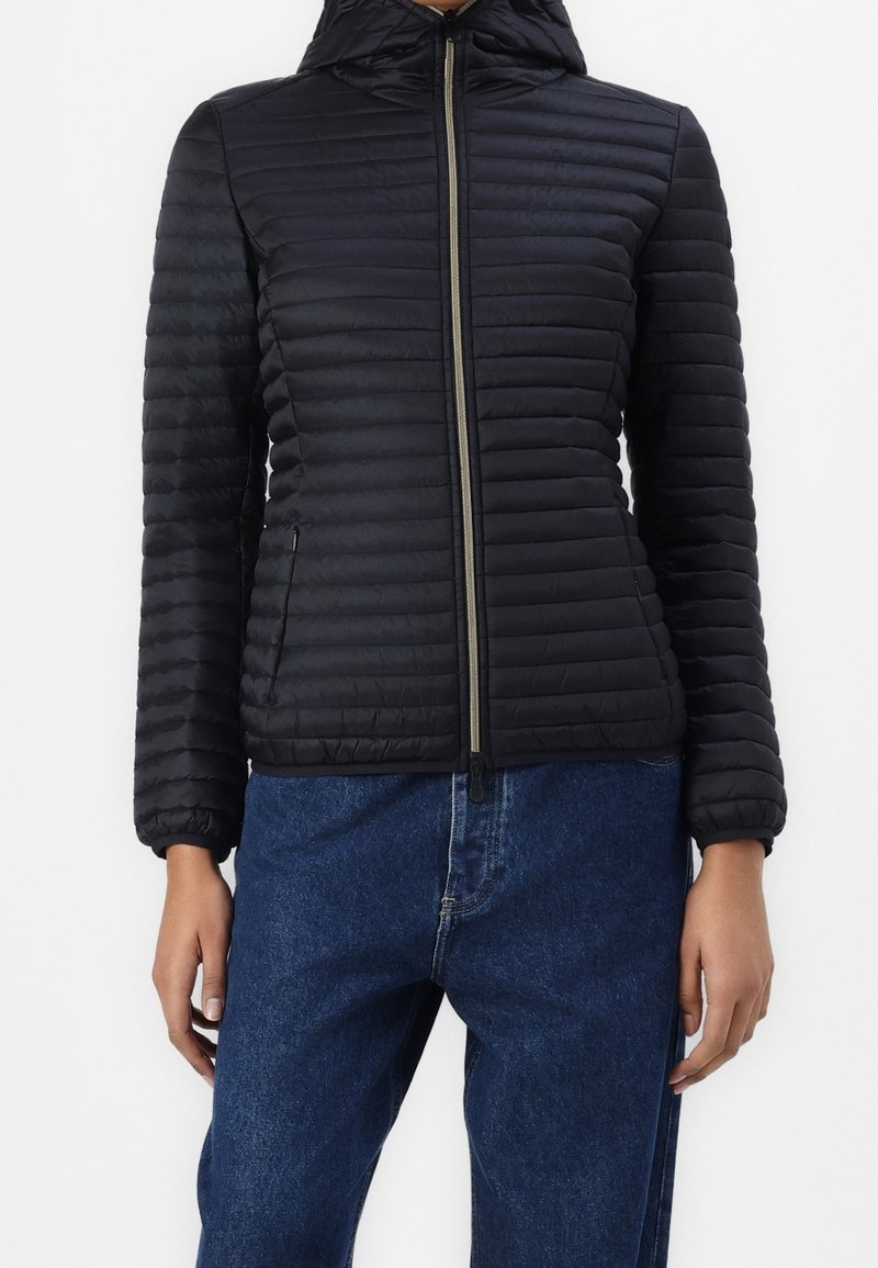 Personne portant une veste matelassée noire à capuche zippée et un jean bleu, debout, les bras détendus le long du corps.