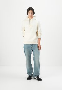 Lichtbeige hoodie met een voorzak en een trekkoord, gecombineerd met vervaagde blauwe jeans. Zwarte sneakers met een textuurdetail maken de look compleet.