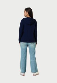 Tengerészkék kapucnis pulóver, bordás mandzsettákkal és aljúval, világoskék, bő szárú farmernadrággal párosítva. Az öltözet laza, kényelmes vágású részletekkel rendelkezik.