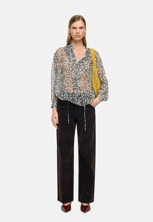 Kobieta w przezroczystej bluzce w kropki z falbanami, czarne spodnie o szerokich nogawkach, brązowe spiczaste buty oraz żółtą teksturowaną torebkę na ramię.