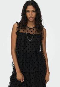 Top nero trasparente con pois, volants sovrapposti e spalline sottili. Presenta una texture morbida e una silhouette elegante.