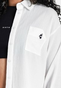 Hvid, struktureret skjorte med knapper og en enkelt brystlomme. Har et sort broderet design på lommen. Sort ribbet undertrøje synlig.