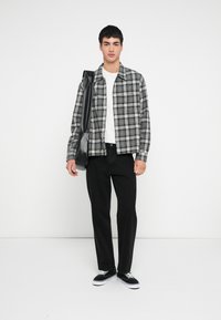 Harmaa-musta ruudullinen paita valkoisen t-paidan päällä, mustat farkut ja mustat lenkkarit valkoisilla pohjilla. Pitelee mustaa olkalaukkua.