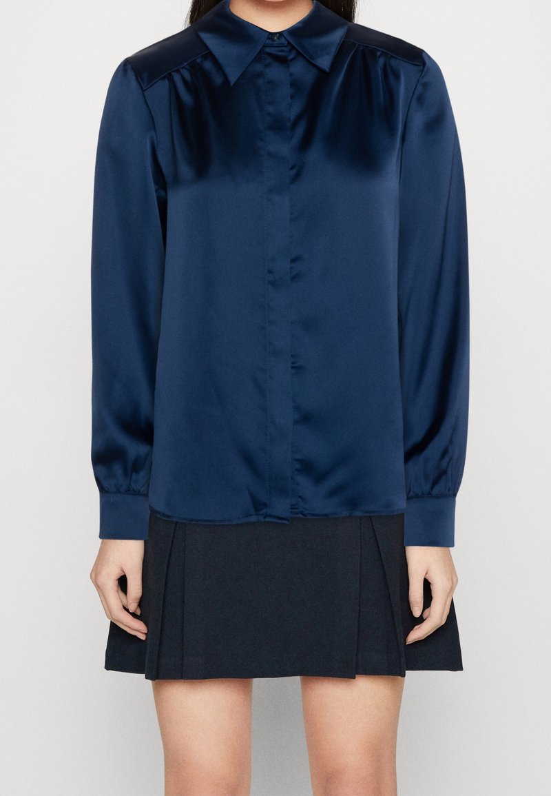 Chemise longue en satin bleu marine à manches longues avec un col pointu, fermeture à boutons sur le devant et des détails de manches froncées, associée à une jupe noire plissée.