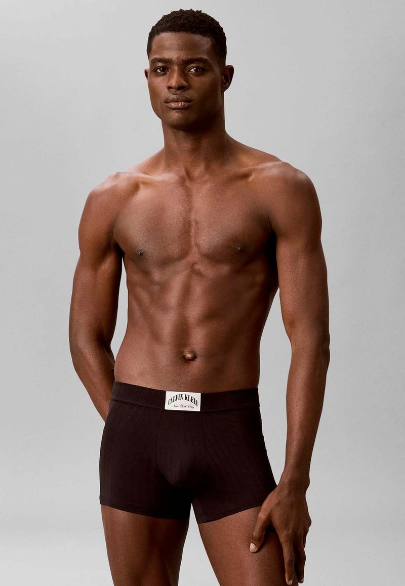 Homme musclé debout devant un fond uni, portant un boxer noir Calvin Klein, regardant devant avec une expression neutre.