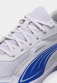 Λευκό αθλητικό παπούτσι με επάνω μέρος από δίχτυ, λευκά κορδόνια, μπλε γραφικό στο πλάι και κείμενο «STABILITY SKELETON» κοντά στην περιοχή των κορδονιών.