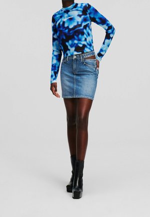 Person iført en blå og sort mønstret langærmet top, denim mininederdel og sorte plateau ankelstøvler mod en ensfarvet baggrund.