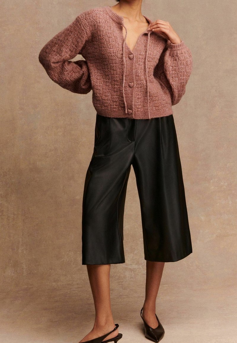 Femme portant un cardigan rose poussiéreux texturé avec des boutons en forme de cœur, un pantalon en cuir noir large et court, et des talons slingback noirs à bout pointu.