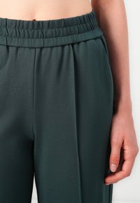 Pantalon vert foncé avec une taille élastique, en tissu lisse, coupe décontractée et plis subtils sur le devant.