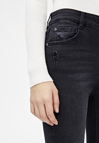 Los jeans de denim negro tienen un diseño ajustado con detalles desgastados en el bolsillo delantero. La cintura tiene un corte clásico, con herrajes plateados.