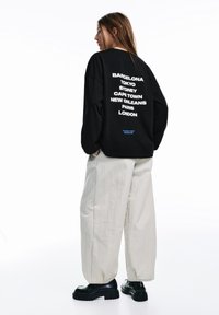 Hanorac negru cu text alb listând nume de orașe pe spate. Asortat cu pantaloni largi, de culoare deschisă, și încălțăminte neagră cu platformă.