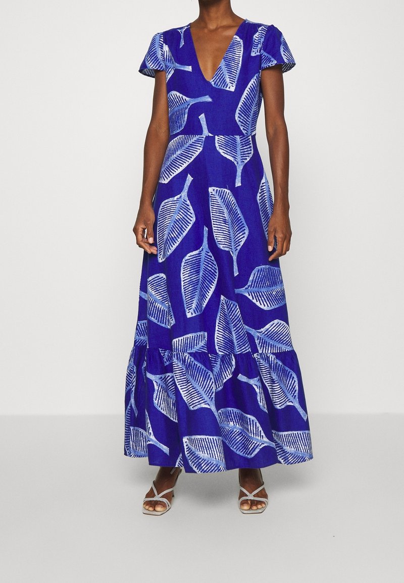 Maxiklänning i klart blått tyg med vita bladmönster, korta volangärmar, djup V-ringning och en flerlagers fåll. Remsandaler i vitt.