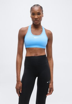 Αθλητική γυναίκα με πλεξούδες, που φορά ανοιχτό μπλε αθλητικό σουτιέν και μαύρο ψηλόμεσο κολάν, στέκεται μπροστά σε απλό φόντο.