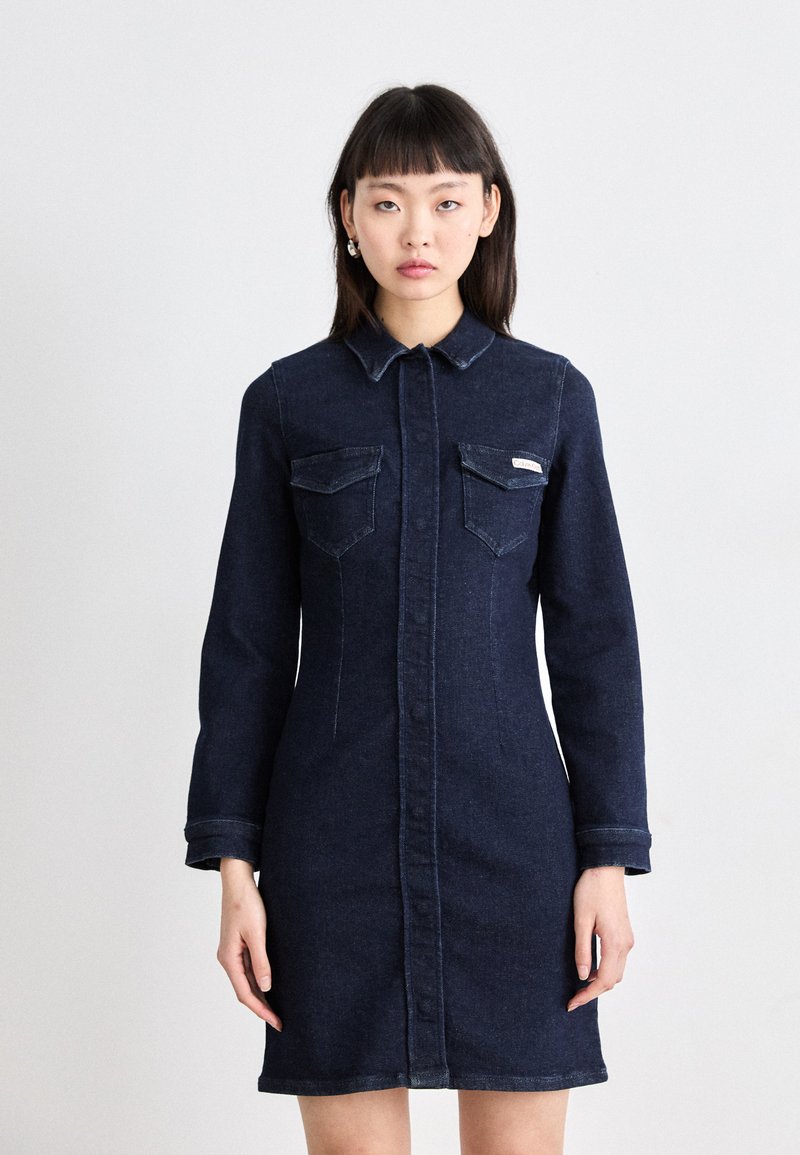 Mørk denimkjole med lange ermer, knappelukking foran, to brystlommer og krage. Har synlige sømmer og en slim fit.