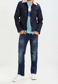 Giacca di jeans con collo color crema, t-shirt grafica blu e jeans blu sbiaditi. Abbinata a sneaker bianche, in piedi contro uno sfondo neutro.