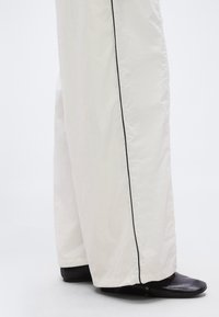 Κάτω άκρα που φορούν λευκό, φαρδύ παντελόνι με μαύρο περιθώριο και μαύρα επίπεδα παπούτσια σε λευκό, απλό φόντο.