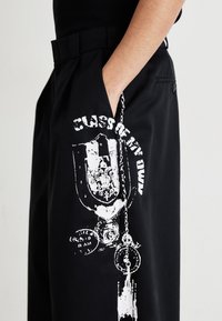 Pantaloni negri cu un buzunar lateral prezintă un design grafic alb proeminent, care încorporează lanțuri și un motif de leu, realizat dintr-o țesătură texturată.