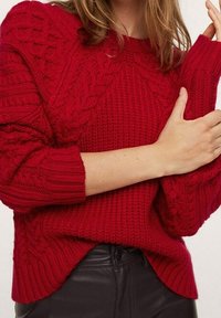 Maglione rosso lavorato a maglia con un motivo testurizzato, caratterizzato da dettagli a trecce e polsini a coste. Vestibilità ampia e design con orlo asimmetrico.