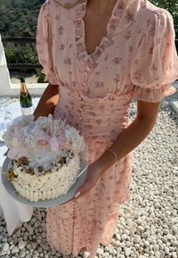 Femme en robe fleurie rose tenant un gâteau blanc décoré surmonté de fleurs, debout sur des cailloux blancs près d'une table avec des boissons.