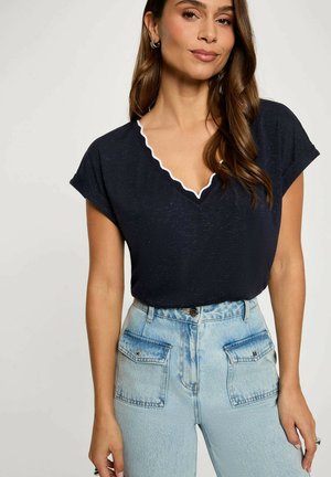 Donna con capelli castani lunghi che indossa una maglietta nera a maniche corte con scollo a V e bordo bianco, abbinata a jeans azzurri a vita alta con tasche sul davanti con pattina.