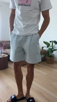 Homme portant un t-shirt graphique blanc, un short gris clair et des sandales noires, debout dans un salon avec un sol en bois et des plantes d'intérieur.
