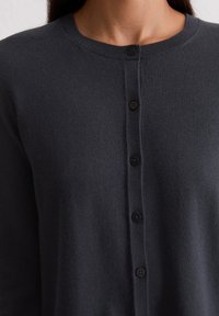 Tuvplāna attēls par personu, kas valkā tumši pelēku adītu džemperi ar piecām redzamām melnām pogām priekšpusē.