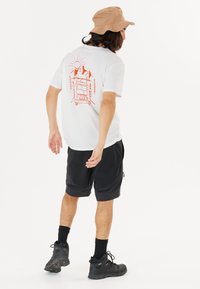Vit T-shirt med en orange grafik av en skåpbil, berg och en sol. Modellen bär svarta shorts och en beige bucket hat, samt svarta skor.