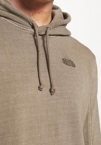 Felpa con cappuccio in morbido tessuto marrone, caratterizzata da un logo testurizzato, polsini a coste e lacci con estremità annodate. Design morbido e casual.