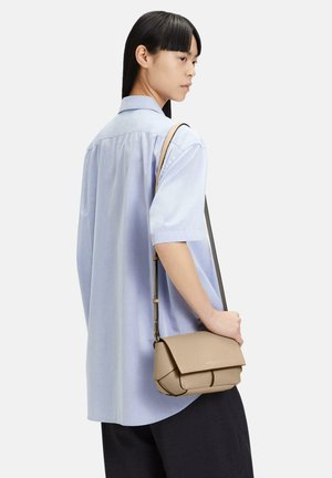 Beige textured leather handbag with a flap closure and adjustable strap, shown hanging from a shoulder of a person in a light blue shirt.