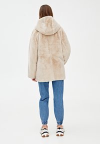 Beige fluffy hættejakke med lange ærmer, baglænsk sømdetaljer, iført blå joggingbukser og hvide sneakers. Blød tekstur er synlig.