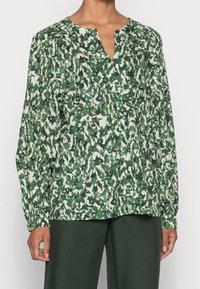 Femme portant une blouse blanche à manches longues avec un motif dense de feuilles vert foncé et un pantalon vert foncé, se tenant devant un fond uni.