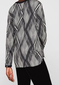 Camicia a maniche lunghe a fantasia geometrica in bianco e nero indossata da una persona con una mano visibile, abbinata a pantaloni neri su uno sfondo semplice.
