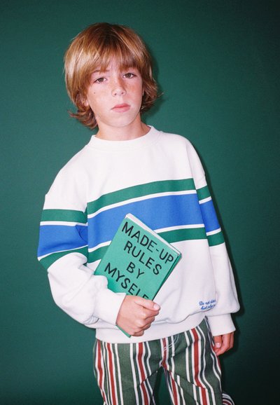 Felpa bianca con strisce blu e verdi, abbinata a pantaloni a righe verdi e rosse. Tiene in mano un libro verde intitolato "REGOLE INVENTATE DA ME."