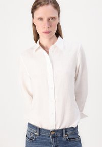 Camicia bianca in lino con colletto classico, maniche lunghe e vestibilità rilassata, abbinata a jeans in denim blu scuro.