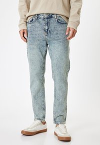 Jeans de un azul claro desgastados, con un corte recto, costuras visibles, cinco bolsillos y cierre de botón, combinados con zapatillas blancas.