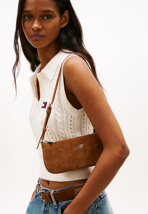 Νέα γυναίκα που φοράει λευκή αμάνικη πλεκτή μπλούζα, μπλε τζιν, καφέ τσάντα χιαστί από σουέτ και καφέ ζώνη με ασημένια πόρπη.