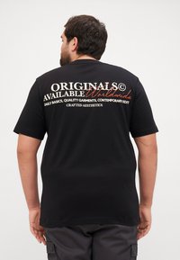 Tricou scurt cu mânecă neagră, având text alb și roșu pe spate. Textul spune „ORIGINALS AVAILABLE Worldwide”, împreună cu detalii suplimentare de brand.