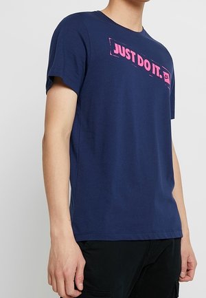 Osoba ubrana w granatowy t-shirt z krótkim rękawem z różowym napisem "JUST DO IT." i logo na piersi, w połączeniu z czarnymi spodniami.