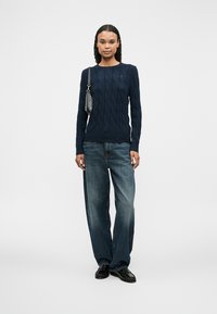 Σκούρο μπλε πουλόβερ με πλέξη καλωδίου και μακριά μανίκια, σε συνδυασμό με φαρδιά μπλε τζιν και μαύρες loafers. Μια άσπρη τσάντα ώμου ολοκληρώνει το σύνολο.