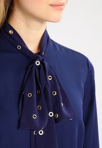 Blouse marine avec un col en nœud comprenant des accents en œillet doré et une texture de tissu lisse et fluide.