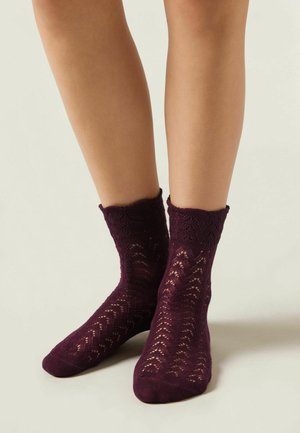 Burgundy bomullsockor till anklarna med ett spetsliknande, texturerat mönster och ribbad kant, vilande på ljus hy mot en neutral bakgrund.