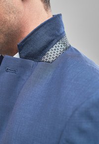 El cuello de una chaqueta azul presenta un detalle texturizado tejido con un patrón geométrico. La tela parece suave con un ligero brillo, destacando los contrastes.