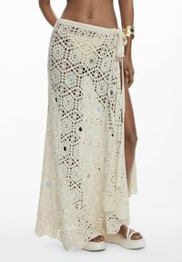 Žena v krémovej maxi sukni z háčkovania s bočným rozparkom, s platformovými sandálmi a zlatým náramkom na zápästí.