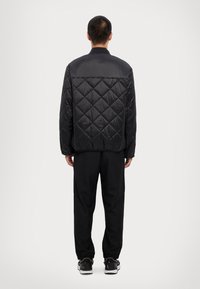 Fekete tűzött pufi dzseki sima gallérral, geometrikus gyémántmintával, valamint fekete nadrággal. A lábbeli kontrasztos fehér talppal rendelkezik.