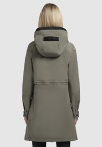 Giacca impermeabile verde oliva con cappuccio, vita regolabile e dettagli neri. Presenta una patch testurizzata con il logo sul retro e polsini con bottoni.